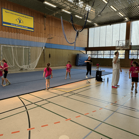 Sechs Kinder in pinken T-Shirts üben in einer Sporthalle, während zwei Erwachsene sie anleiten.