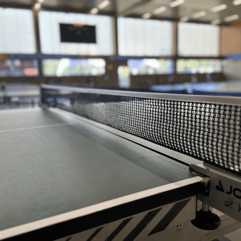 Nahaufnahme einer grünen Tischtennisplatte mit Netz und Teilansicht der Sporthalle im Hintergrund.