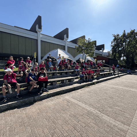 Gruppe von Kindern in pinken T-Shirts sitzt auf Steinstufen vor einem Gebäude an einem sonnigen Tag.
