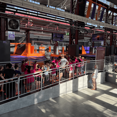 Gruppe von Kindern in pinken T-Shirts steht an einer Trampolinfl&auml;che, w&auml;hrend ein Trainer Anweisungen gibt.