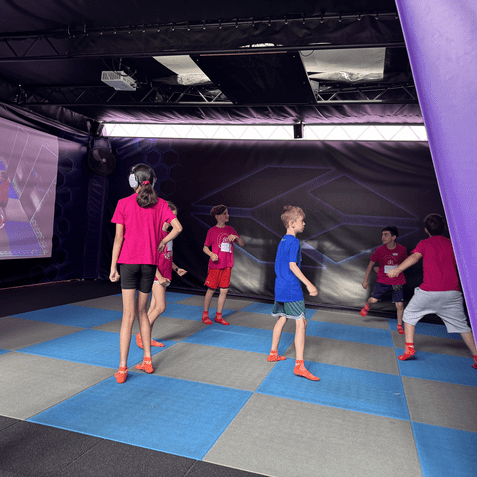 Gruppe von Kindern in bunten T-Shirts spielt in einer Indoor-Arena mit interaktiven Projektionen auf einem Spielboden.
