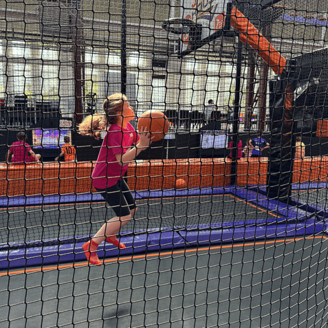 M&auml;dchen springt auf einem Trampolin und wirft einen Basketball zum Korb in einer Trampolinhalle.