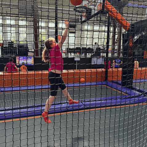 M&auml;dchen springt in einem Trampolinpark zum Basketballkorb, um einen Ball zu werfen, umgeben von Netzen.