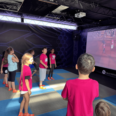 Gruppe von Kindern in pinken Shirts steht vor einer Projektionsfl&auml;che und interagiert mit einem Spiel.