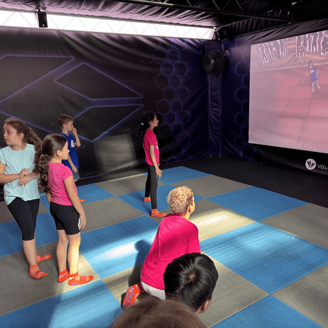 Gruppe von Kindern in bunten T-Shirts schaut auf einen Bildschirm mit interaktivem Spiel in einem Freizeitbereich.