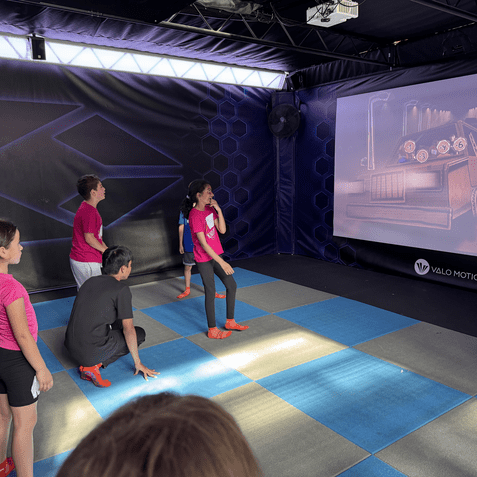 Gruppe von Kindern interagiert mit einem Projektionsspiel auf einer farbigen Spielmatte in einem Indoor-Spielbereich.