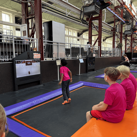 Kinder in pinken T-Shirts sitzen an einer Trampolinfl&auml;che, ein Kind springt auf dem Trampolin vor einem Videodisplay.