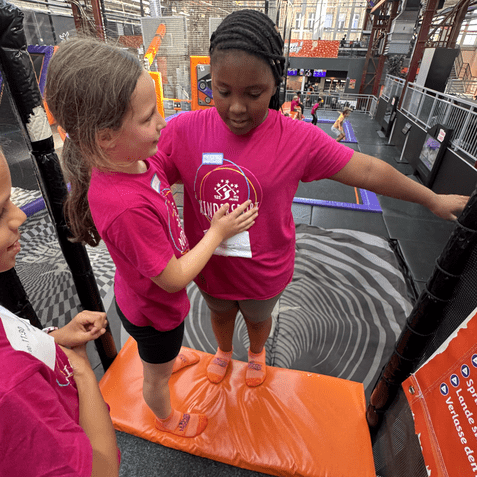 Zwei Kinder in pinken T-Shirts stehen auf einem orangenen Mattenfeld und interagieren in einer Trampolinhalle.