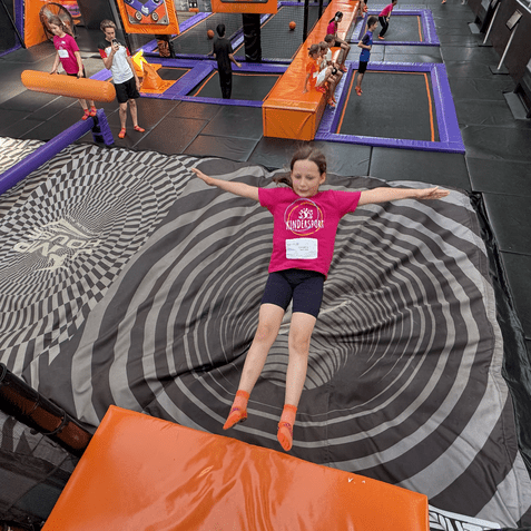 M&auml;dchen in pinkem Shirt springt auf ein gro&szlig;es, spiralf&ouml;rmiges Sprungtuch in einem Trampolinpark.
