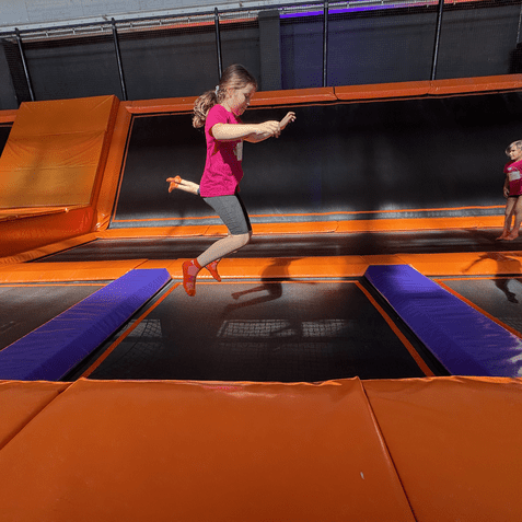 Drei Kinder springen auf Trampolinen in einer Trampolinhalle mit orangefarbenen und blauen Elementen.