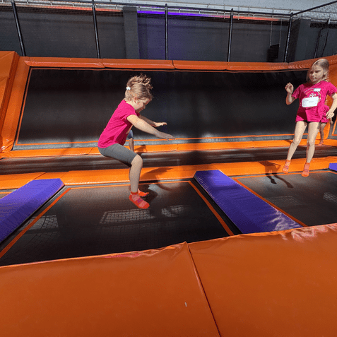 Zwei M&auml;dchen in pinken T-Shirts springen auf Trampolinen in einer Trampolinhalle mit orangefarbenen Elementen.