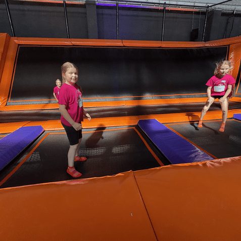 Kinder springen auf Trampolinen in einer Trampolinhalle mit orangefarbenen und lila Unterlagen.