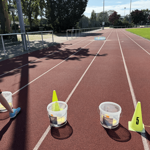 Eine Person in kurzer Sportbekleidung geht auf einer Laufbahn vorbei an Beh&auml;ltern und Markierungskegeln.