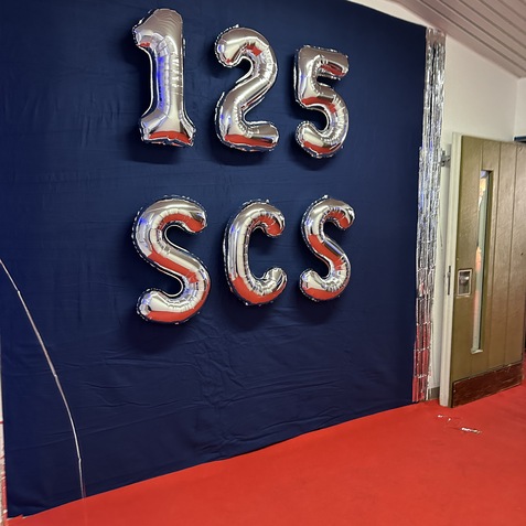 Silberne Ballons mit den Zahlen &bdquo;125&ldquo; und dem Buchstaben &bdquo;SCS&ldquo; h&auml;ngen vor einem blauen Hintergrund an einer rotem Teppich.