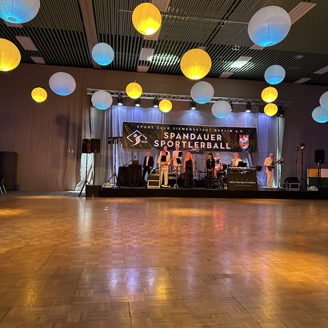Beleuchteter Festsaal mit MusikerInnen auf der B&uuml;hne und h&auml;ngenden Papierlaternen in verschiedenen Farben.