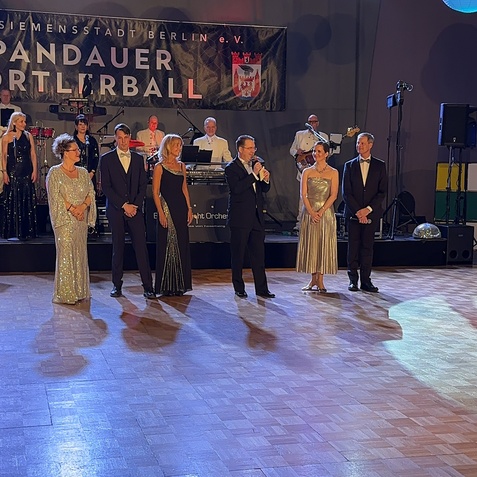 Gruppenbild von G&auml;sten in eleganter Kleidung beim Pandauer Sportball, mit einer Band im Hintergrund und bunten Laternen.