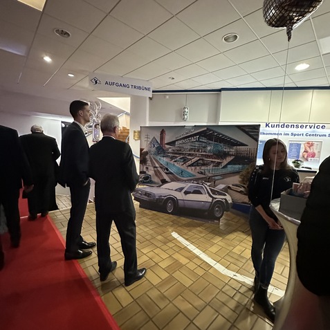 Menschen stehen an einem Informationsstand in einer Veranstaltungshalle, im Hintergrund ein Banner mit einem Sportzentrum.