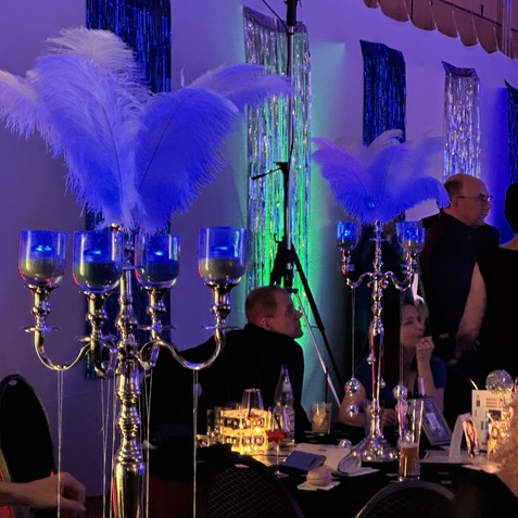 Dekorative Tischleuchte mit Federn und blauen Kerzen auf festlich gedecktem Tisch bei einer Veranstaltung. Tischgäste im Hintergrund.