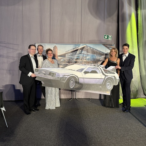 Fünf Personen in Abendgarderobe stehen vor einem großen Poster eines DeLorean-Fahrzeugs in festlicher Atmosphäre.