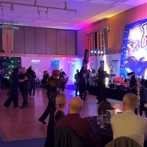 Tanzende Paare und G&auml;ste an Tischen in einer festlich beleuchteten Halle mit buntem Hintergrund.