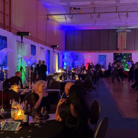 Festliche Veranstaltung in einem beleuchteten Saal mit Tischen, Gästen und farbiger Dekoration an den Wänden.