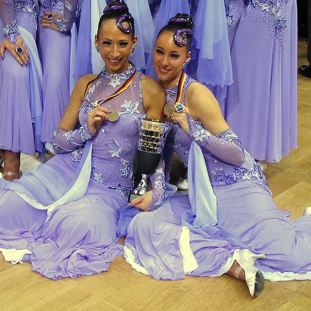 Zwei Tänzerinnen in lila Kostümen sitzen lächelnd auf dem Boden und halten einen Pokal, umringt von weiteren Tänzerinnen.