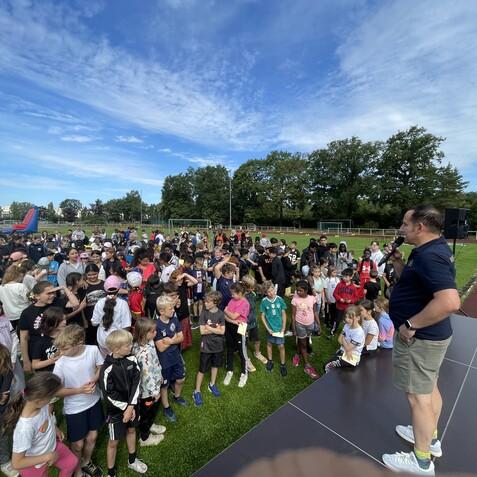Eine Gruppe von Kindern versammelt sich auf einem Sportplatz, w&auml;hrend ein Mann auf einer B&uuml;hne spricht.