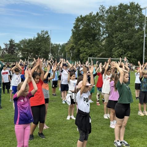 Gruppentraining von Kindern im Freien, die mit erhobenen H&auml;nden ein &Uuml;bungsgel&auml;nde auf einer Wiese absolvieren.