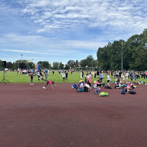Gruppierung von Kindern und Jugendlichen bei einem Sportevent auf einer Wiese, umgeben von Rucksäcken und einer Bühne.