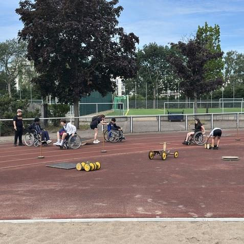 Gruppen von Jugendlichen in Rollstühlen trainieren auf einer Sportanlage mit verschiedenen Trainingsgeräten und Hindernissen.