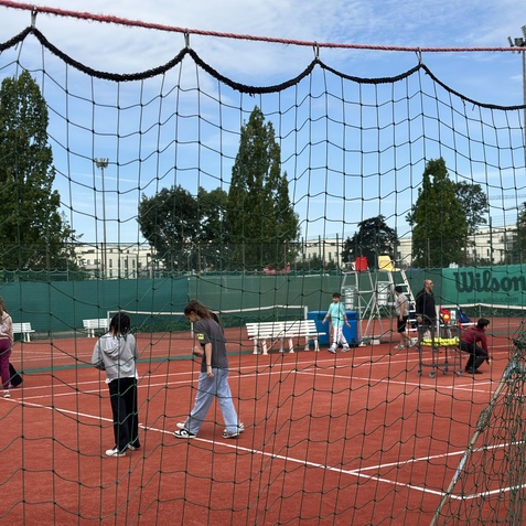 Sechs Kinder und Erwachsene spielen Tennis auf einem roten Platz, umgeben von Bäumen und einer Netzumrandung.