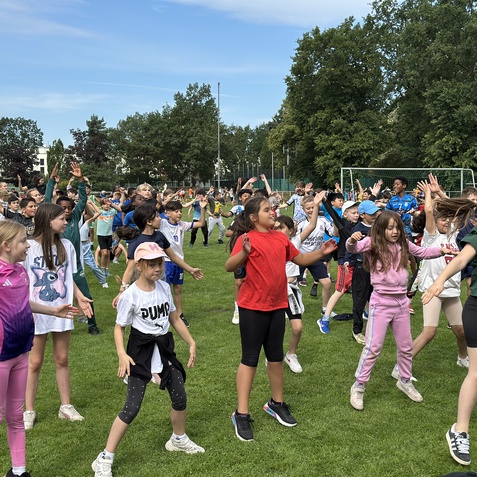 Gruppierung von Kindern in sportlicher Aktivität auf einer Wiese, viele mit erhobenen Armen und verschiedenen Outfits.