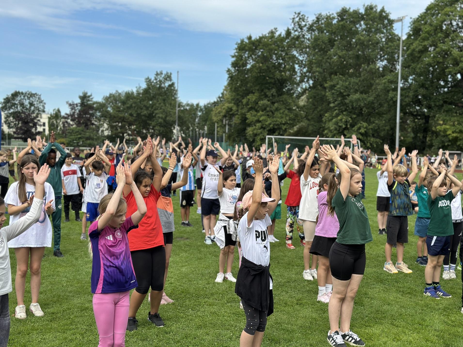Gruppentraining von Kindern im Freien, die mit erhobenen H&auml;nden ein &Uuml;bungsgel&auml;nde auf einer Wiese absolvieren.