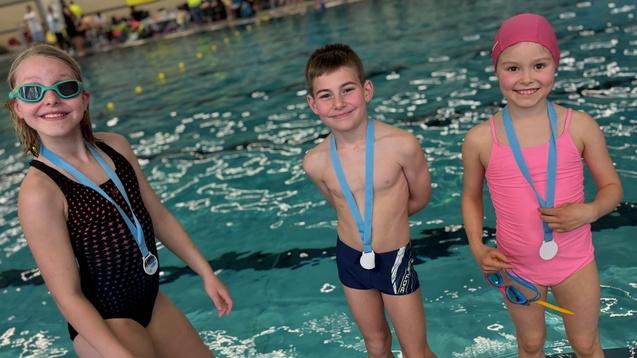 Drei Kinder in Schwimmbekleidung mit Medaillen stehen am Rand eines Schwimmbeckens und lächeln in die Kamera.
