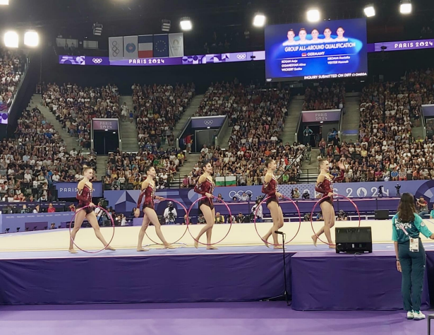 Sechs Turnerinnen in roten Kostümen führen eine Gymnastikübung mit Reifen vor einem Publikum in der Sportarena aus.
