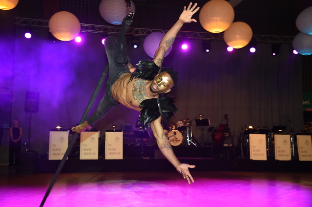 Akrobat in Federkostüm schwebt an einem Seil über eine Bühne mit Musikerensemble und bunten Lampions im Hintergrund.