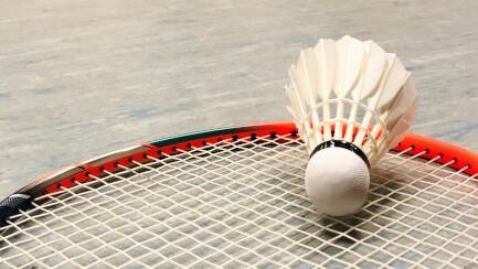 Nahaufnahme eines Badmintonschlägers mit Federball auf dem Boden einer Sporthalle im Hintergrund Spieler.
