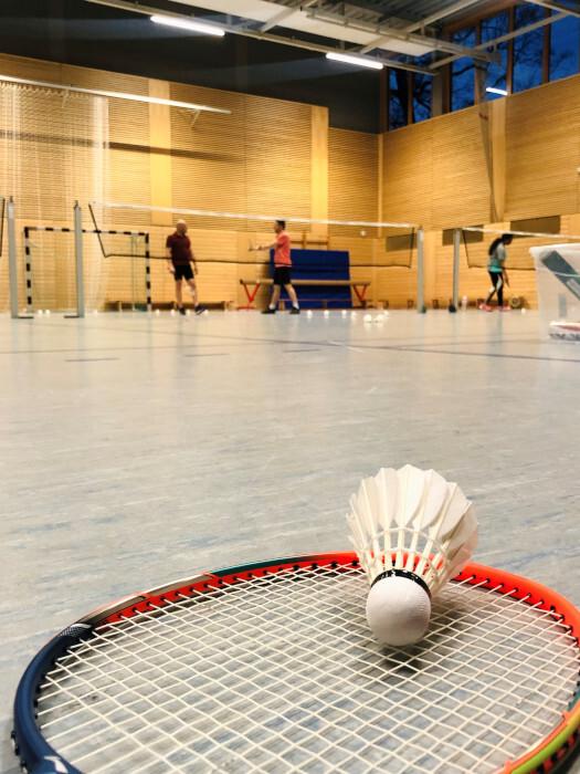Nahaufnahme eines Badmintonschlägers mit Federball auf dem Boden einer Sporthalle im Hintergrund Spieler.