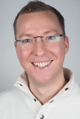 L&auml;chelnder Mann mit Brille in einem wei&szlig;en Pullover vor einem hellen Hintergrund.