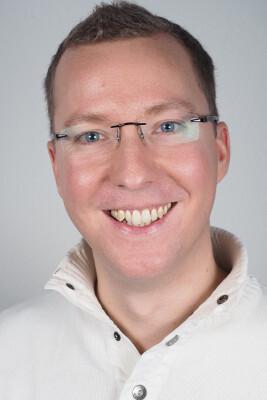 Lächelnder Mann mit Brille in einem weißen Pullover vor einem hellen Hintergrund.