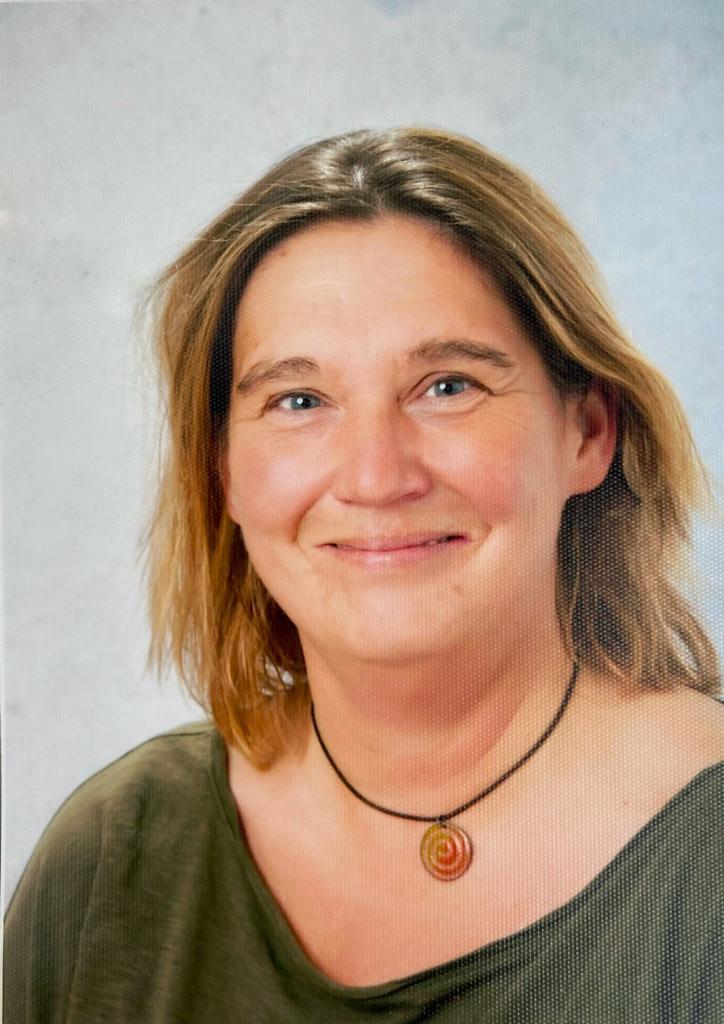 Porträt einer lächelnden Frau mit blonden, leicht unordentlichen Haaren und einem schwarzen Halsband mit einer roten Perle.