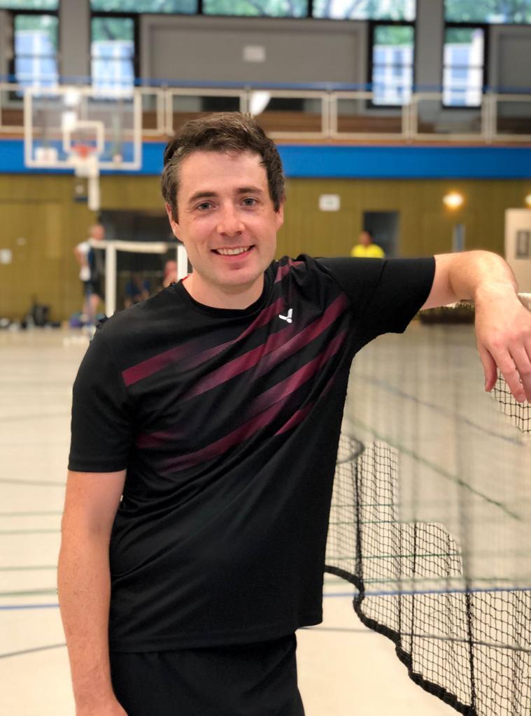 Mann in schwarzem Sportshirt lehnt entspannt an einem Tischtennisschläger bei einer Sportveranstaltung in einer Halle.