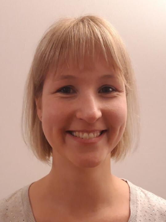 Lächelnde Frau mit blonden Haaren, die in einem hellen Pullover vor einer neutralen Wand steht.
