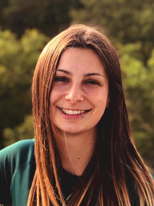 Porträt einer lächelnden jungen Frau mit langen, braunen Haaren und Piercing, vor unscharfem, grünem Hintergrund.