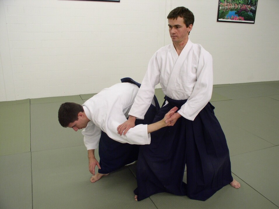 Zwei Kampfsportler üben eine Technik auf einer Matte, einer beugt sich nach vorne, während der andere ihn hält.