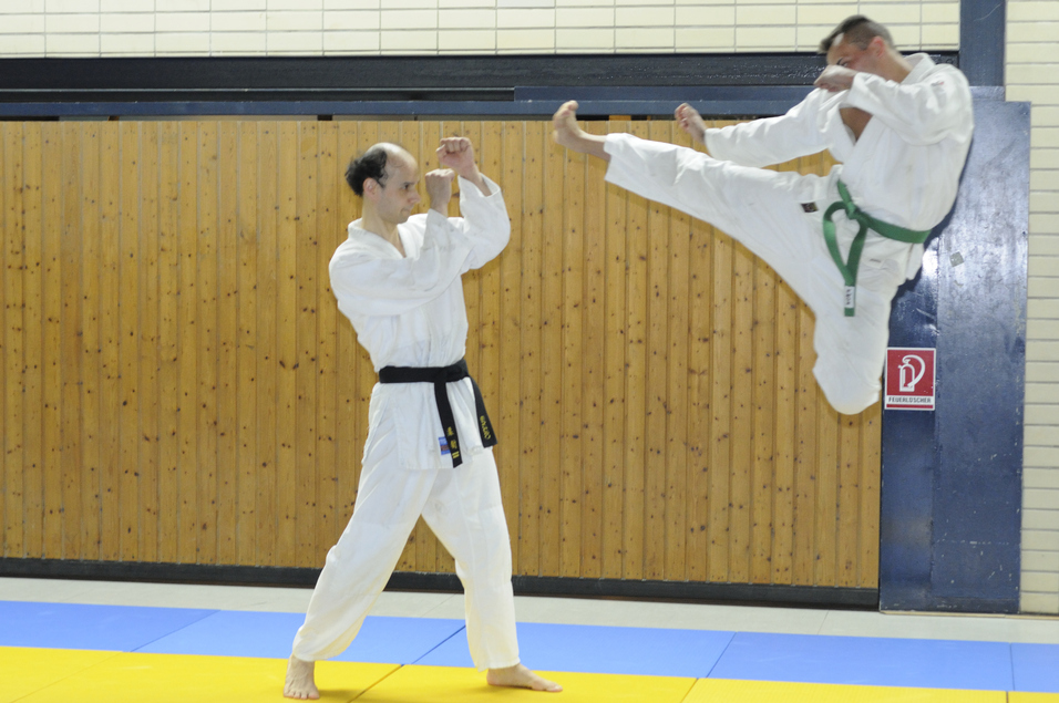 Zwei Martial-Arts-Praktizierende in wei&szlig;en Anz&uuml;gen, einer f&uuml;hrt einen hohen Kick aus, der andere ist bereit.