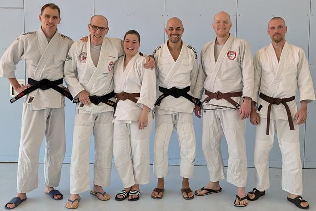 Gruppe von sechs Kampfsportlern in Gi, zwei mit schwarzen, vier mit braunen Gürteln, stehen vor einer blauen Wand.