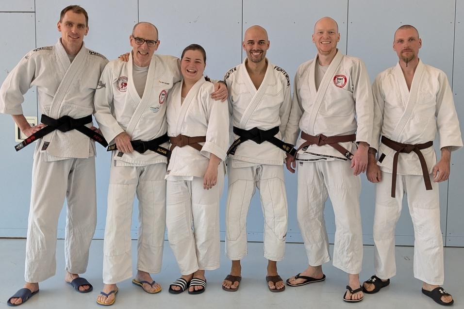 Gruppe von sechs Kampfsportlern in Gi, zwei mit schwarzen, vier mit braunen G&uuml;rteln, stehen vor einer blauen Wand.