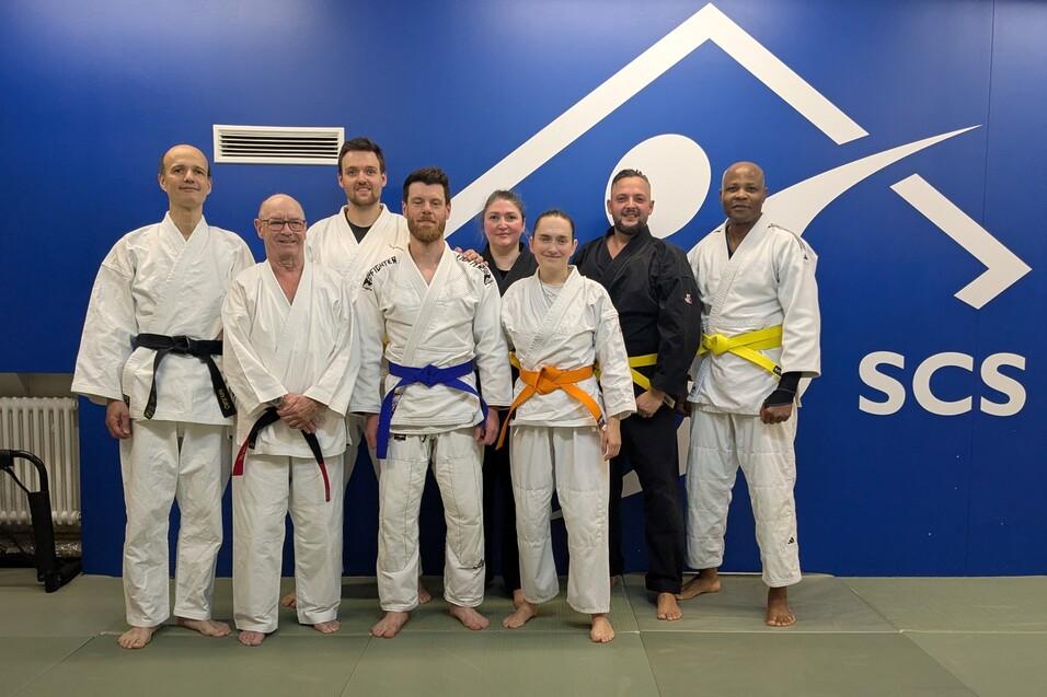 Gruppe von acht Personen in Judokleidung mit verschiedenen G&uuml;rtelfarben vor einer blauen Wand des SCS-Studios.