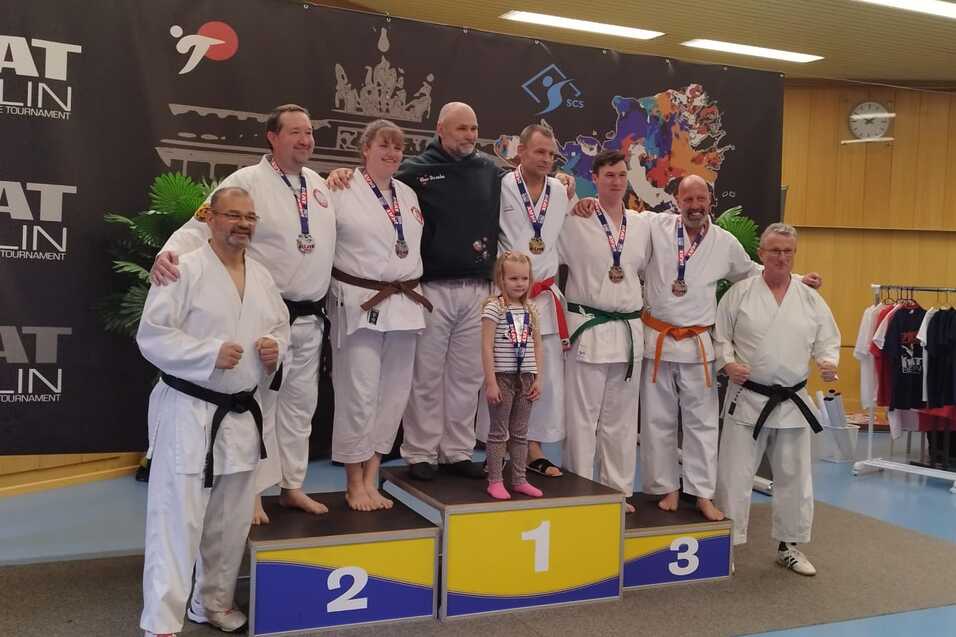 Sieben Karateka in Uniform stehen auf einem Podium mit Medaillen, umgeben von einer dekorativen Kulisse.